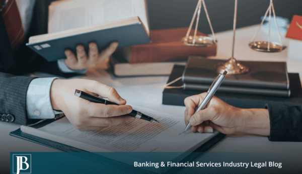 Two business attorneys seated at a desk reviewing and signing a commercial collection litigation defense agreement, with law books and gold scales of justice in the background, on the Jimerson Birr Banking and Financial Services Industry Legal Blog.