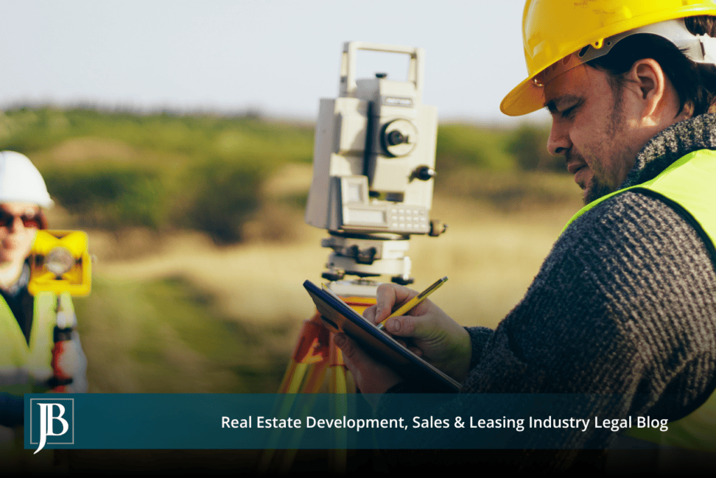 Two surveyors in hard hats and reflective vests using a total station and taking notes along a grassy right-of-way corridor, illustrating eminent domain in Escambia County road projects.