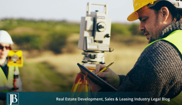 Two surveyors in hard hats and reflective vests using a total station and taking notes along a grassy right-of-way corridor, illustrating eminent domain in Escambia County road projects.