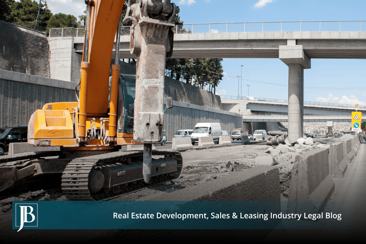 Excavator with a hydraulic breaker demolishing a concrete median barrier along an active interstate widening project beneath an elevated overpass.