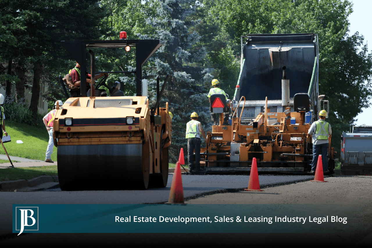 Road construction crew paving a two-lane roadway with an asphalt paver and steel-drum roller behind orange traffic cones, with workers in high-visibility vests directing the operation.