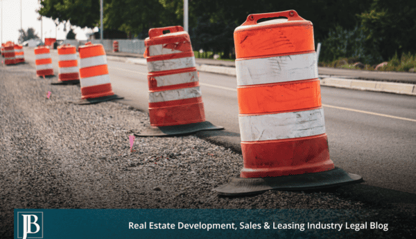Orange and white traffic barrels and pink survey flags lining the gravel shoulder of a two-lane road under widening construction.
