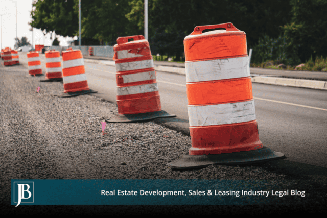 Orange and white traffic barrels and pink survey flags lining the gravel shoulder of a two-lane road under widening construction.