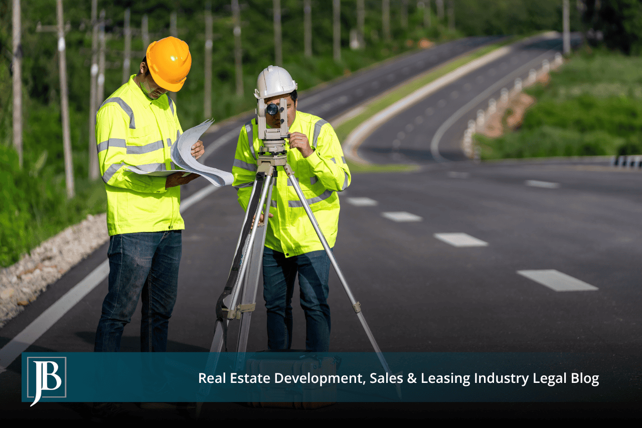 Two surveyors in hard hats and high-visibility yellow jackets stand on a newly paved multi-lane road, one sighting through a tripod-mounted survey instrument while the other reviews a set of plans.