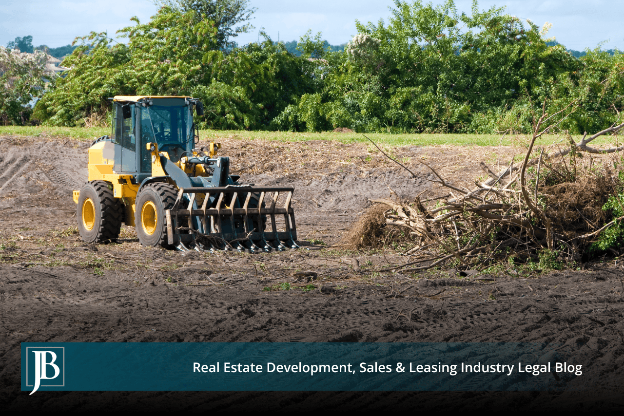 Yellow wheel loader with a grapple rake attachment clearing brush from a freshly graded dirt lot, beside a pile of uprooted branches and a green tree line