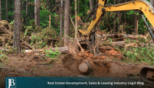 Excavator clearing felled trees and stumps from a wooded parcel during highway right-of-way construction