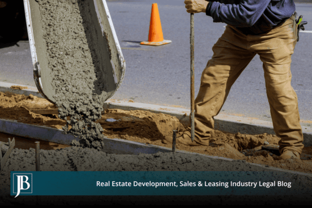Wet concrete pours from a truck chute into a new curb form along a roadway while a construction worker levels the mix with a hand tool beside an orange traffic cone.