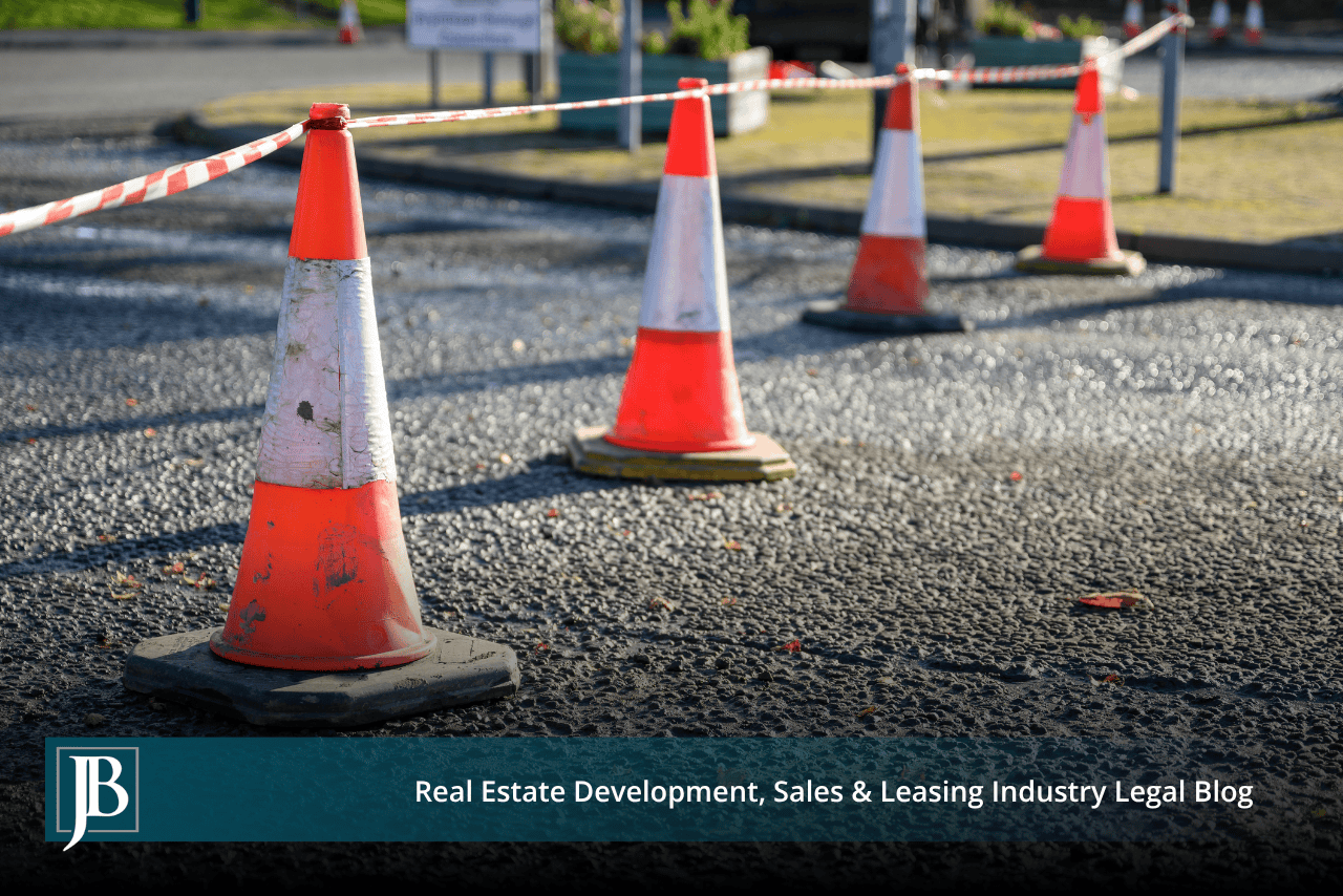 Orange and white traffic cones joined by red and white barrier tape on freshly paved asphalt, with a grass median and planters in the background