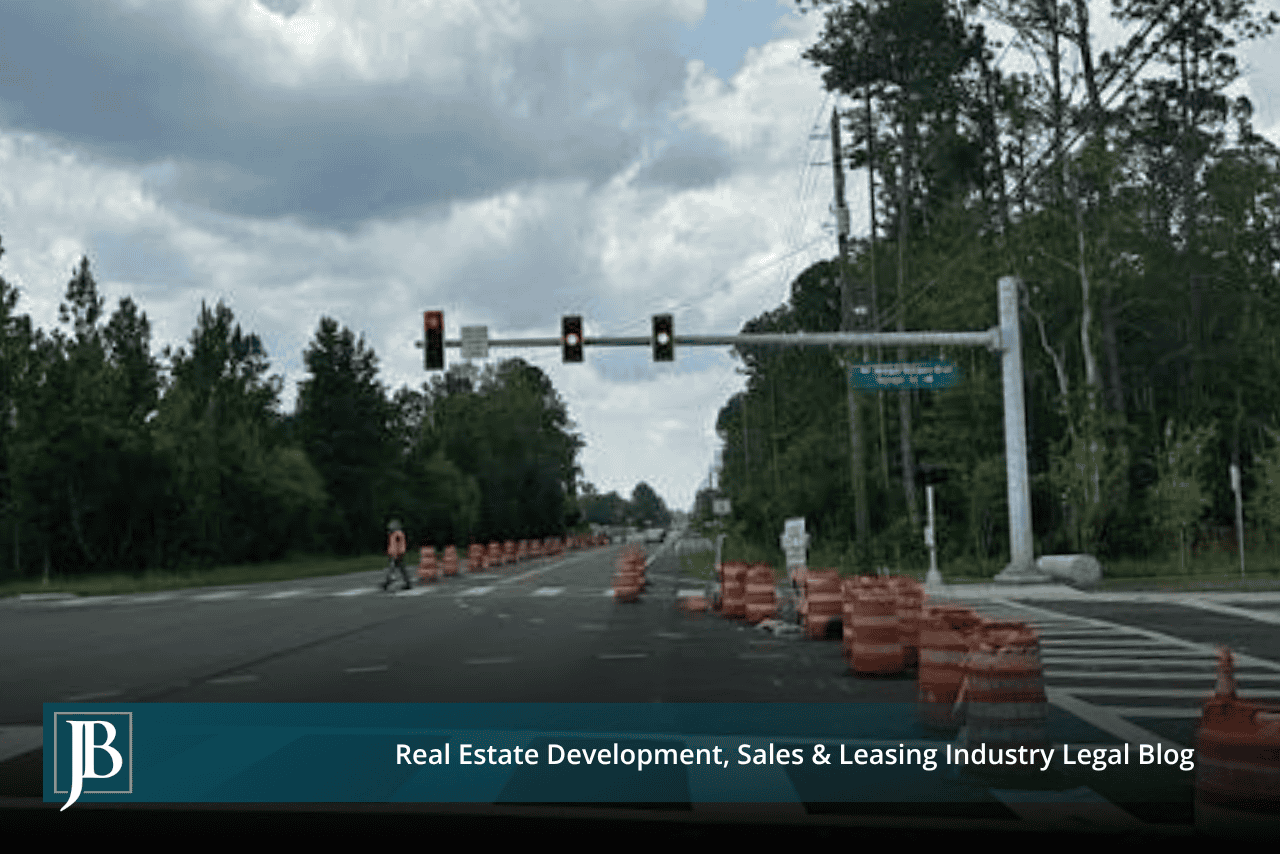 Orange construction barrels line a newly signalized highway intersection bordered by tall pines, with a worker crossing the road beneath a green street sign on the signal mast arm